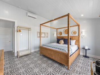 A bedroom with a four-poster bed, patterned floor tiles, and modern decor, featuring wall art and a small side table by a window.