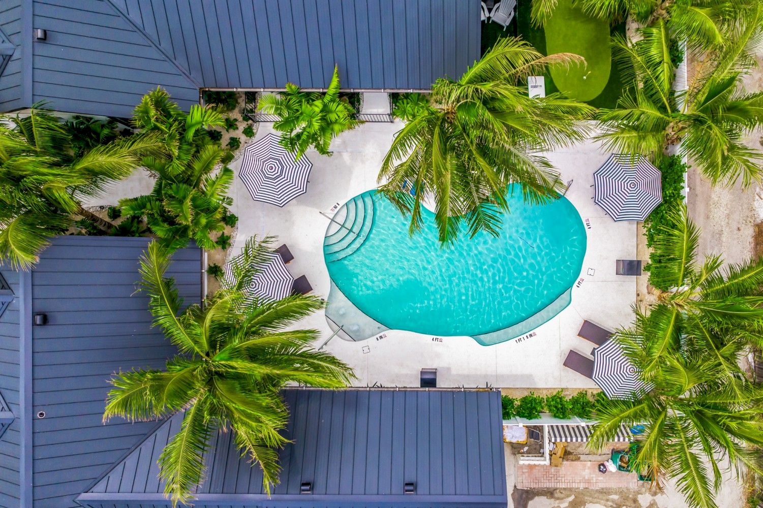 An aerial view of a kidney-shaped pool surrounded by palm trees, lounge chairs, and umbrellas, enclosed by gray-roofed buildings.
