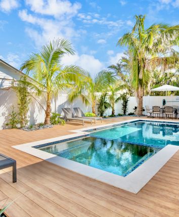A backyard features a modern pool with a wooden deck, surrounded by palm trees, lounge chairs, and a patio dining area.