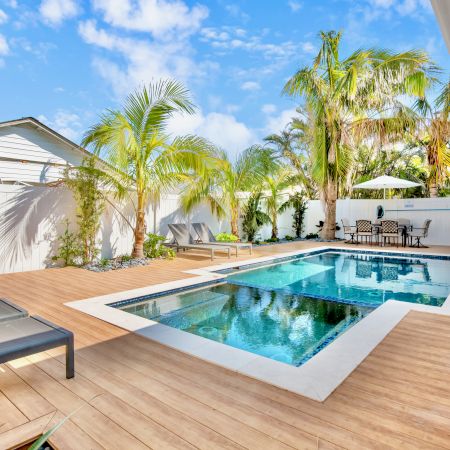 A backyard features a modern pool with a wooden deck, surrounded by palm trees, lounge chairs, and a patio dining area.