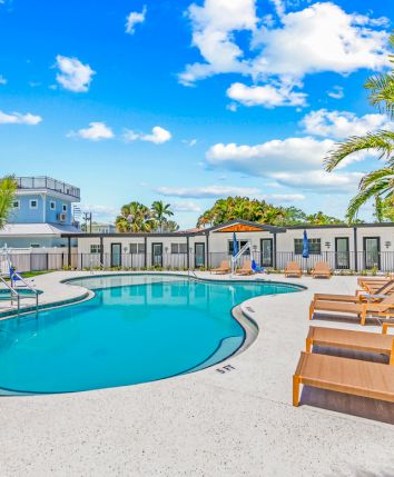 The image shows a sunny pool area with lounge chairs, umbrellas, and palm trees, set against a backdrop of a clear blue sky and clouds.