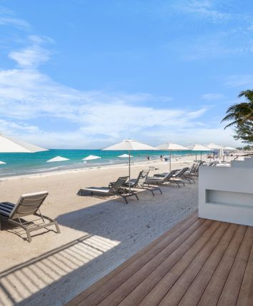 Beach scene with lounge chairs, umbrellas, a wooden deck, and palm trees under a clear blue sky, adjacent to a building.