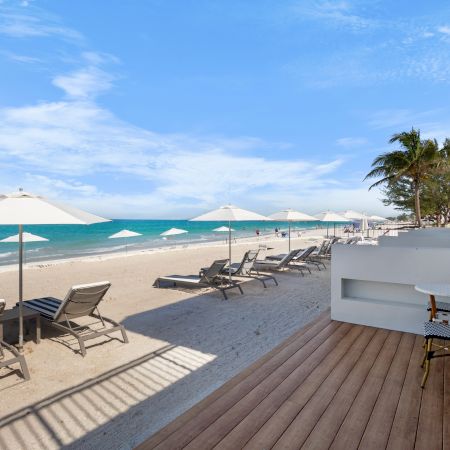 Beach scene with lounge chairs, umbrellas, a wooden deck, and palm trees under a clear blue sky, adjacent to a building.