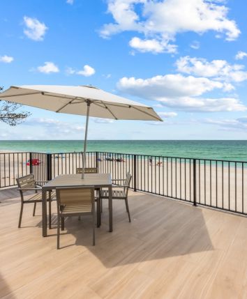 A beachfront deck with a table, chairs, and an umbrella overlooks the sandy beach and ocean under a blue sky with clouds.