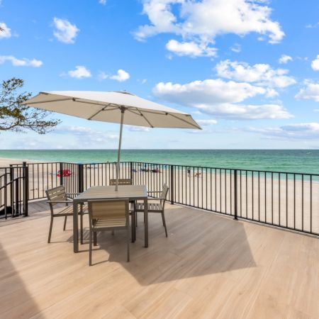 A beachfront deck with a table, chairs, and an umbrella overlooks the sandy beach and ocean under a blue sky with clouds.