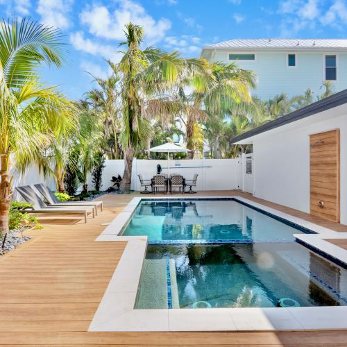 A modern backyard with a swimming pool, palm trees, lounge chairs, and a wooden deck, surrounded by a white fence and a house.
