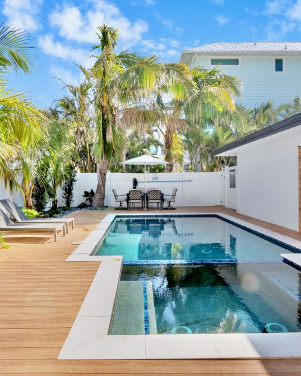 A modern backyard with a swimming pool, palm trees, lounge chairs, and a wooden deck, surrounded by a white fence and a house.