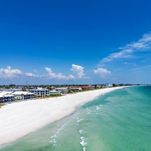 A scenic beach with clear turquoise water, white sand, and seaside houses under a bright blue sky with clouds in the distance.
