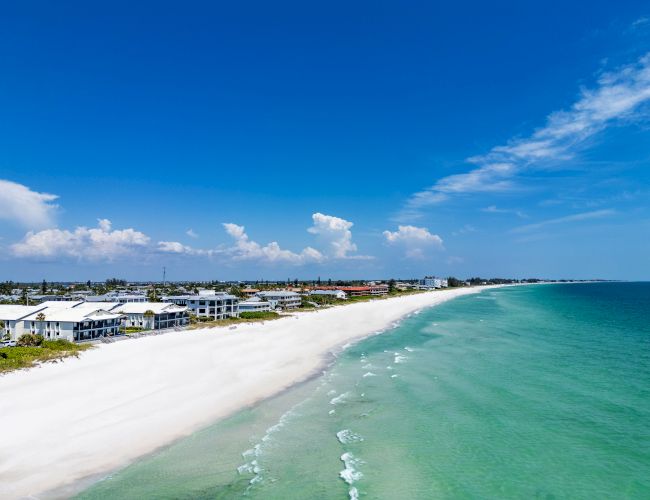 A scenic beach with clear turquoise water, white sand, and seaside houses under a bright blue sky with clouds in the distance.