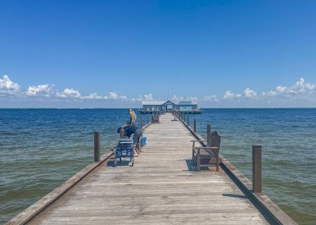 A person relaxes on a long wooden pier extending into a vast body of water, under a clear blue sky with scattered clouds, ending at a structure.