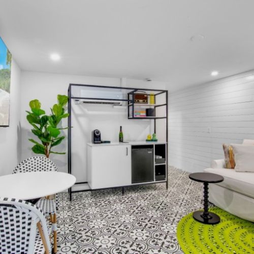 A modern room with a TV, plants, white furniture, small tables, patterned floor, and colorful wall art in a bright, open space.