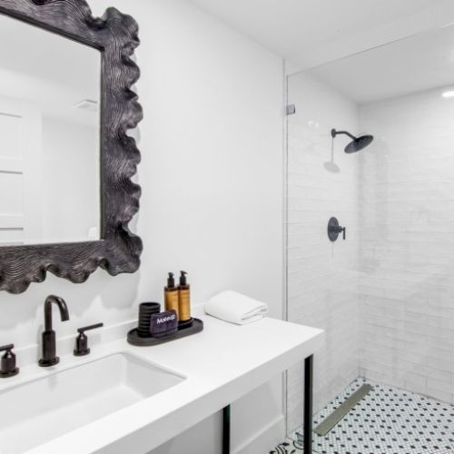 The image shows a modern bathroom with a white sink, decorative mirror, black fixtures, toiletries on the counter, and a glass-enclosed shower.