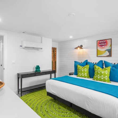 A modern bedroom with a white bed, blue and green pillows, a green rug, wall art, and wicker basket decor on the table.