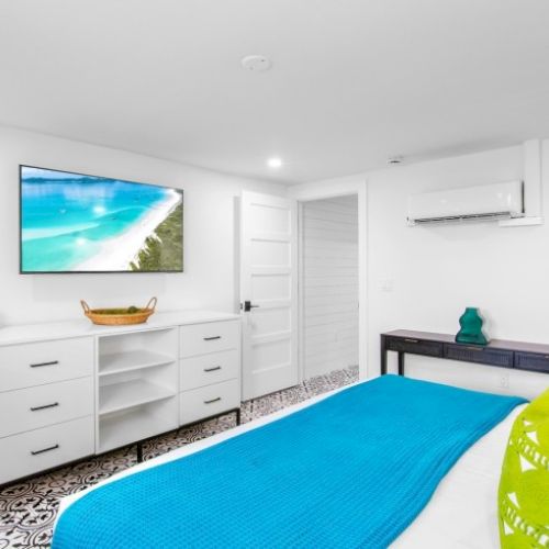 A bright bedroom features a bed with blue bedding, a wall-mounted TV, white furniture, and decorative accents for a serene atmosphere.