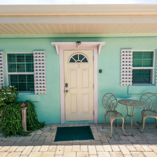 A pastel-colored building with a door, two windows, plants, and a small outdoor metal table and chairs set.