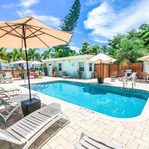 A sunny pool area with lounge chairs, umbrellas, and tropical greenery, featuring a small building and clear sky in the background.