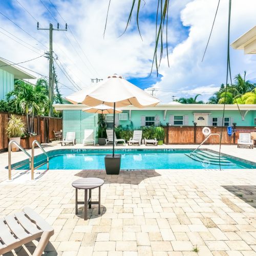 A sunny poolside area with lounge chairs, umbrella, and lush greenery in a fenced yard.