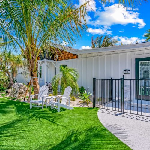 A modern house with a fenced entry, white exterior, palm trees, green artificial turf, and outdoor seating under a blue sky.