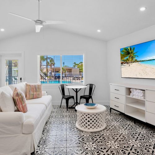 A bright living room with a white sofa, patterned floor tiles, TV, and a small table set against a window and door.