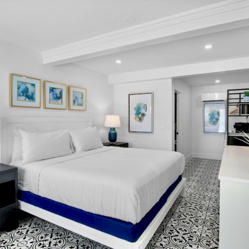 A modern bedroom with a white and blue color scheme, featuring a bed, lamps, artwork, a TV, and a plant on a dresser.