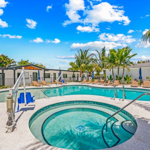 The image shows a pool area with a large pool, a smaller hot tub, palm trees, and lounge chairs under a sunny blue sky.