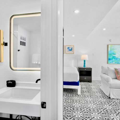 This image shows a stylish room with a bathroom, featuring a sink, mirror, framed art, a bed, sofa, patterned floor tiles, and modern decor.