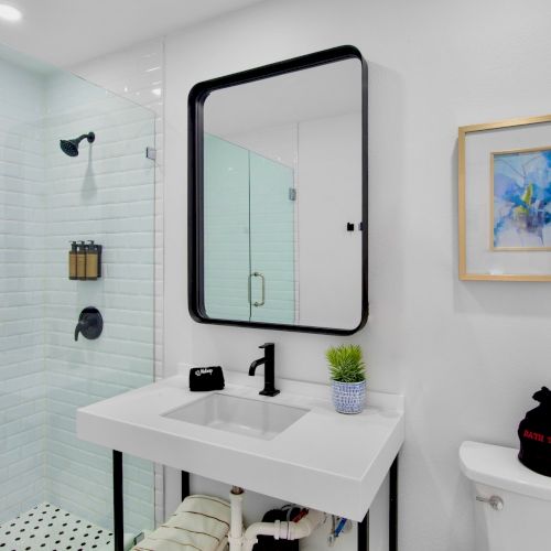 A modern bathroom with a glass shower, white sink, black fixtures, framed art, and a plant. There’s a small item on the toilet.