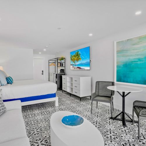 A modern, stylish hotel room features a bed, couch, table with chairs, TV, and vibrant artwork with blue and green tones.