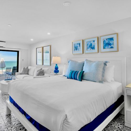 A bright, modern bedroom with a large bed, blue accents, and an ocean view through sliding doors leading to a balcony.