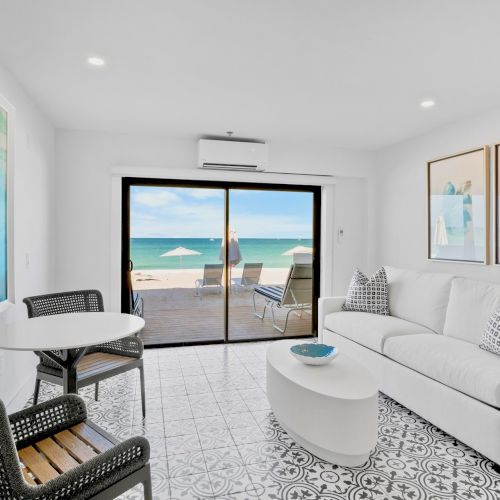 A bright living room with coastal decor, a couch, artwork, and a table, overlooking a beach through sliding glass doors, creating a serene vibe.