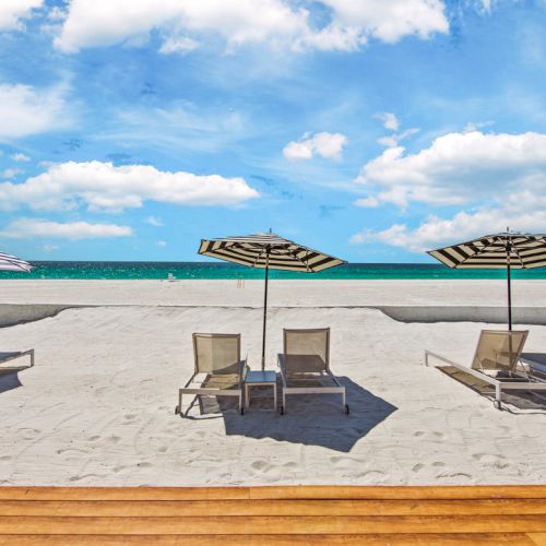 Sandy beach with striped umbrellas and lounge chairs, facing the ocean under a bright blue sky with scattered clouds.