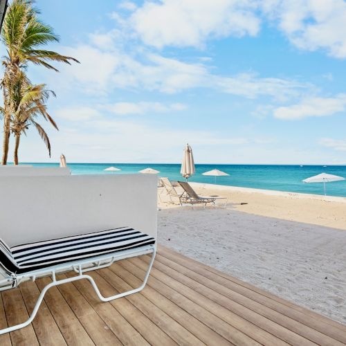 A beachfront view with a striped lounge chair on a deck, umbrellas on the sand, and palm trees under a blue sky.