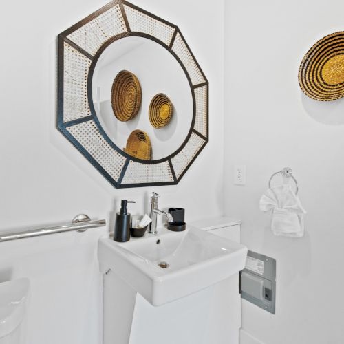 A modern bathroom features a geometric mirror, round wicker wall decor, a small white sink with black and silver dispensers, and a towel ring.