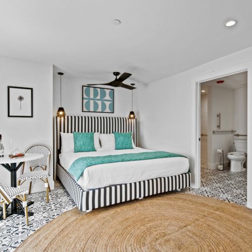 A modern bedroom with a striped bed, turquoise accents, a small table, and an ensuite bathroom with patterned flooring.