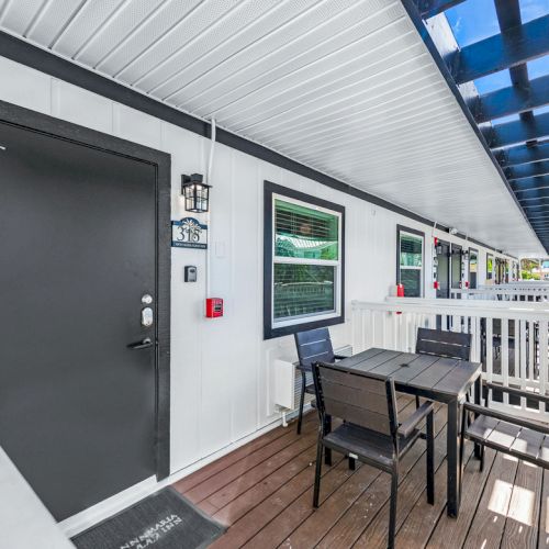 The image shows an outdoor seating area with a dark door, table and chairs on a wooden deck, under a covered walkway with railings.