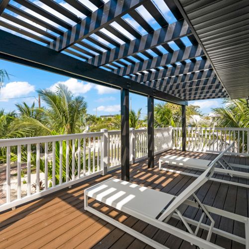 A deck with lounge chairs, white railing, and a pergola, surrounded by palm trees under a clear blue sky.