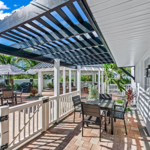 A patio with tables, chairs, a pergola, and outdoor seating on a sunny day in a garden setting with palm trees.