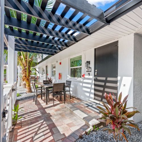 A patio area with outdoor furniture, under a pergola, surrounded by plants and a well-maintained pathway, next to a white building.