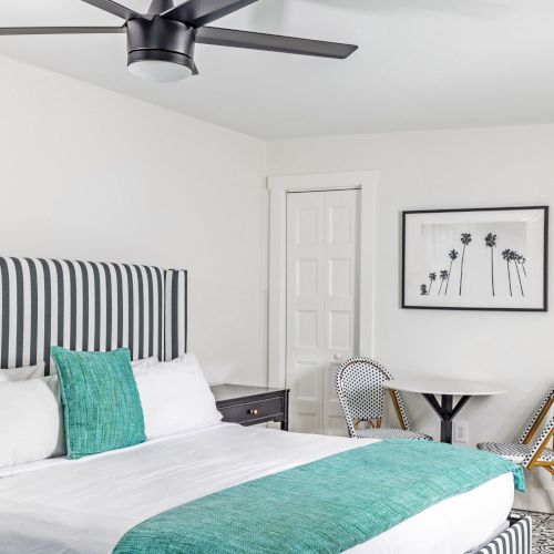The image shows a modern bedroom with a striped headboard, teal bedding, table and chairs, black ceiling fan, and a flower artwork.