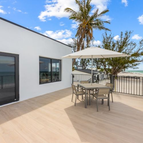 A sunlit patio with a table and umbrella overlooks the ocean, surrounded by railings, a white building, and palm trees under a blue sky.