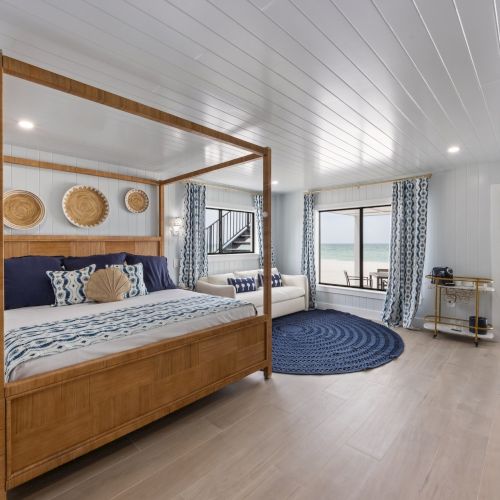 A spacious bedroom with a wooden canopy bed, ocean view windows, blue decor, and a cozy seating area with a round rug.
