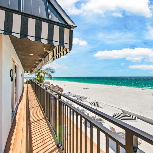 A balcony view shows a sandy beach with striped umbrellas and the ocean under a blue sky. Striped awnings shade the wooden walkway.