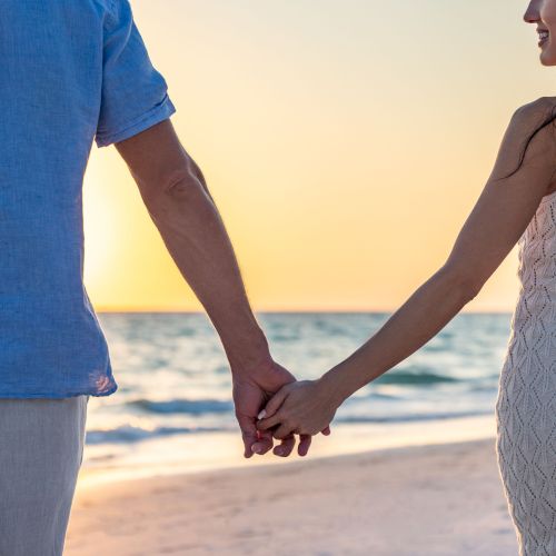 A couple is holding hands while walking on the beach at sunset, with the ocean in the background.