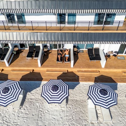 Aerial view of a beachfront property with blue and white umbrellas on sandy beach. Chairs are on the wooden deck near a building.