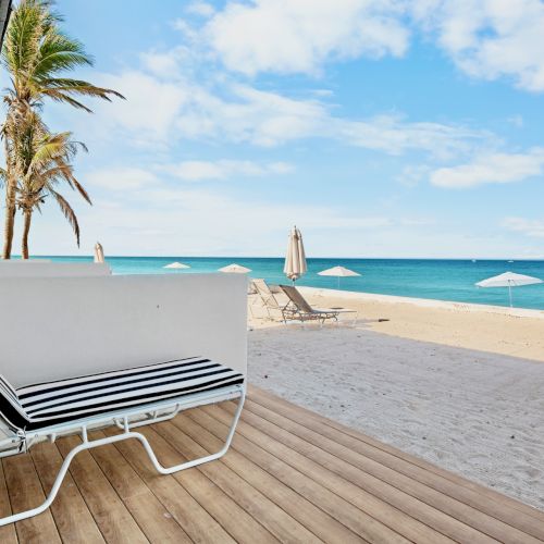 A beachfront scene with lounge chairs, umbrellas, palm trees, and a view of the ocean under a partly cloudy sky.