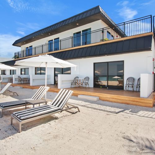 A beachfront building with lounge chairs and umbrellas on the sandy shore, clear blue sky in the background.