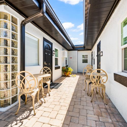A narrow patio with elegant chairs, tables, and a potted plant is nestled between white buildings with black accents, under a bright blue sky.