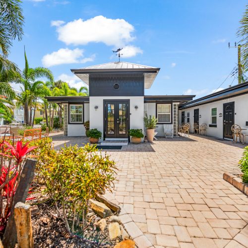 A modern house with a wide driveway, surrounded by tropical plants, blue sky, and outdoor seating; a bright, welcoming atmosphere.
