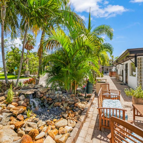 A tropical backyard features palm trees, a rock garden with a small waterfall, and patio seating near a house with large windows.