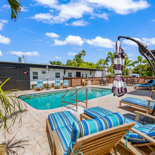 A serene outdoor pool area with lounge chairs, umbrellas, and lush greenery under a bright blue sky with scattered clouds.